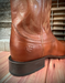 Brown leather boot with embossed Ariat logo on the back heel on a wooden floor