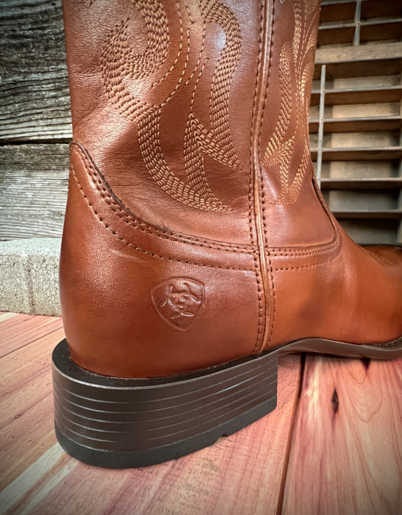 Brown leather boot with embossed Ariat logo on the back heel on a wooden floor