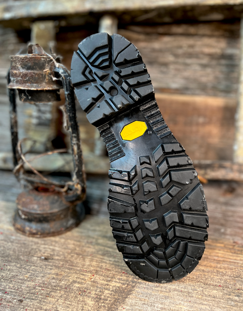 Close-up of a black boot sole with a yellow label on a wooden surface with a lantern in the background.