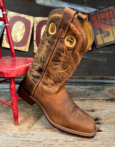 Brown cowboy boot with intricate patterns on a wooden floor with a red chair and rustic wall decorations.