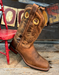 Brown cowboy boot with intricate patterns on a wooden floor with a red chair and rustic wall decorations.