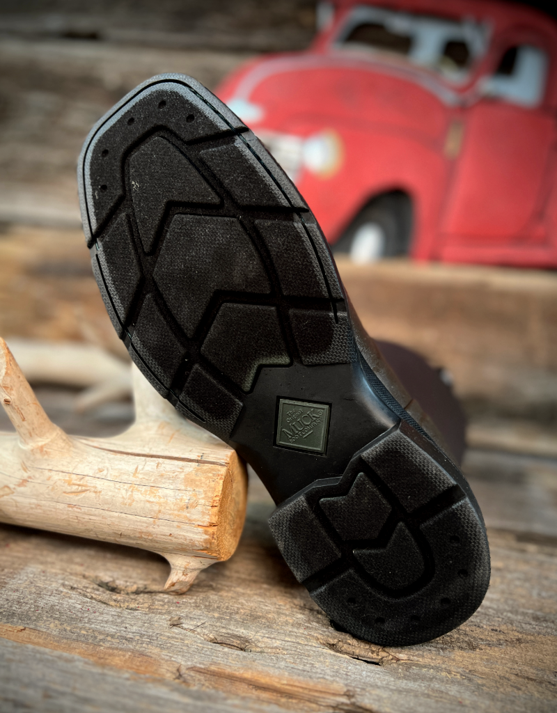 Close-up of a black boot sole on a wooden surface with a red vehicle in the background.