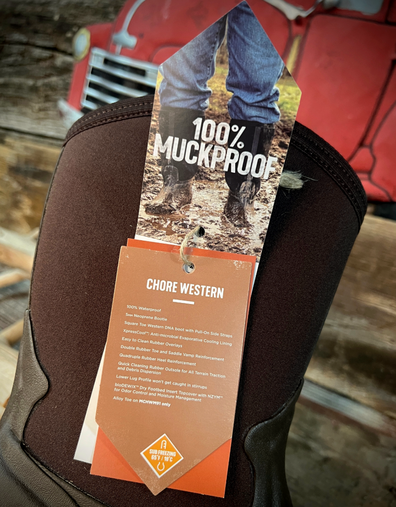 Brown boot with a '100% Muckproof' tag on a wooden surface with a red vehicle in the background.