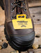 Brown work boot with a yellow 'Steel Toe' tag on a wooden surface with stones.