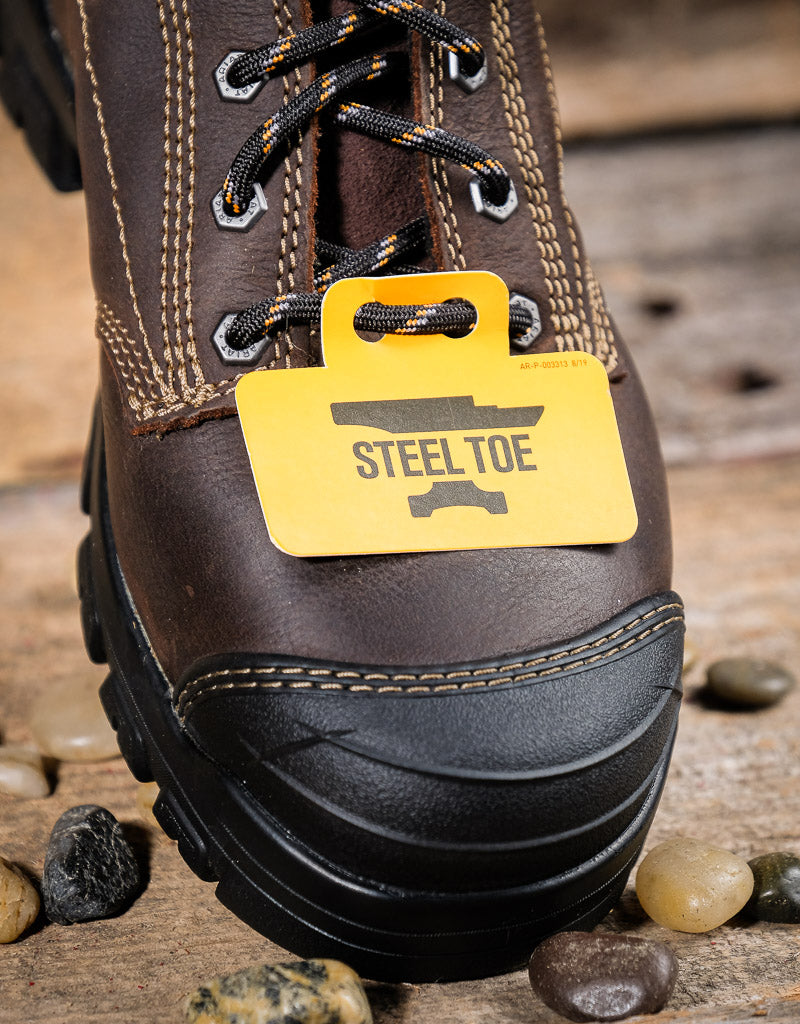 Brown work boot with a yellow 'Steel Toe' tag on a wooden surface with stones.