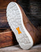 Brown leather shoe with white sole and orange logo on a wooden surface