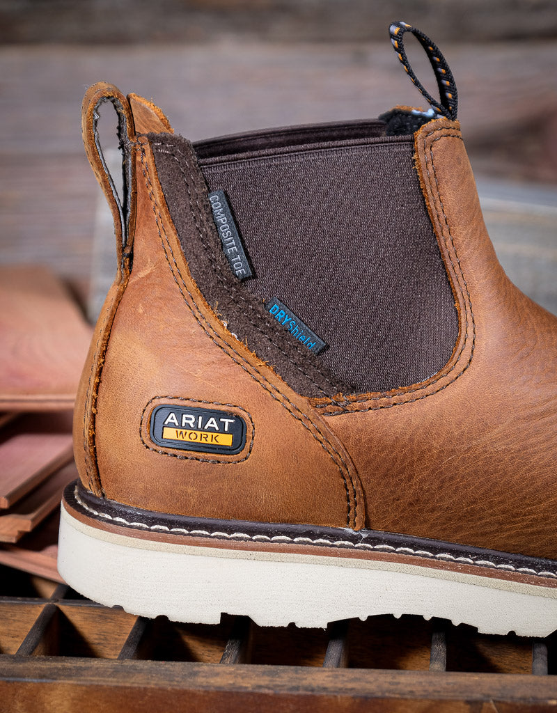 Brown Ariat work boot with a visible brand logo and 'composite toe' and 'DRY shield' tags on a wooden surface.