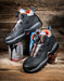 A pair of black safety work boots with BOA dials and orange accents, displayed on a rustic wooden background with paint cans as props.