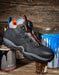 A dark gray and black safety work boot with orange and light blue accents, featuring a BOA dial closure system. The boot is styled on a rustic wooden surface with paint cans in the background.