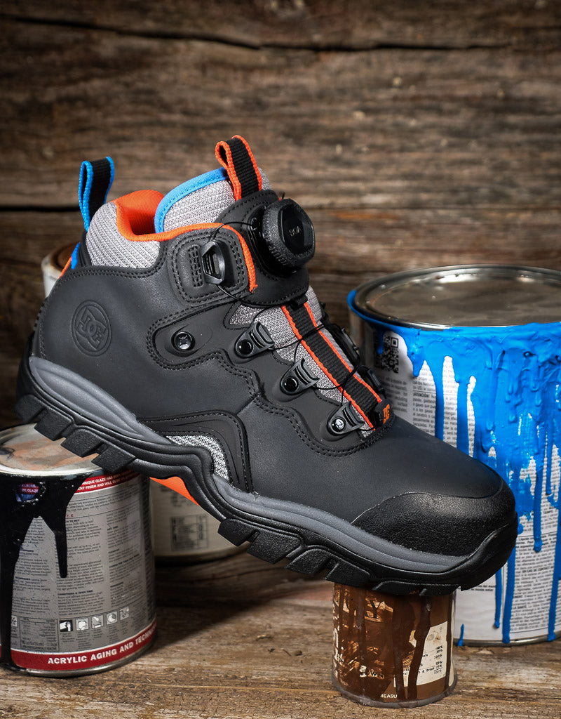 A dark gray and black safety work boot with orange and light blue accents, featuring a BOA dial closure system. The boot is styled on a rustic wooden surface with paint cans in the background.