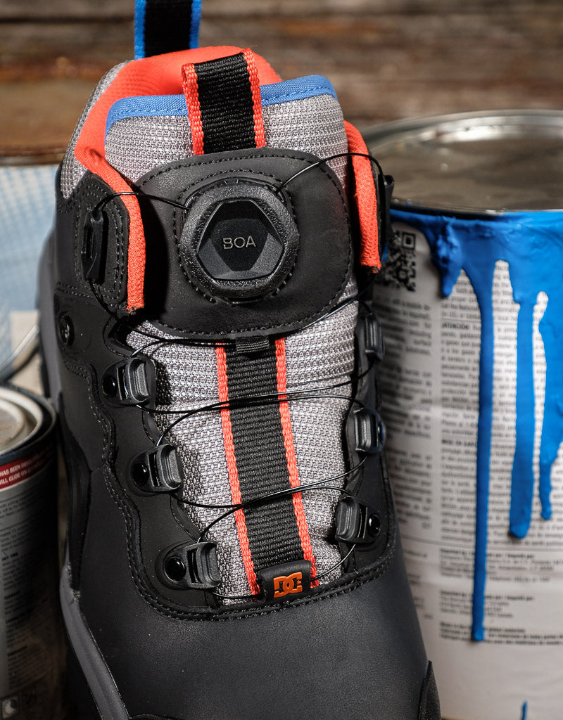 A close-up of a black DC boot with gray mesh panels, bright orange accents, and a BOA dial closure, positioned next to paint cans on a rustic wooden surface.