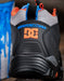 Close-up of the heel of a DC Shoes sneaker in black, gray, and orange, with blue 'DCSHOECOUSA' text on the side. 