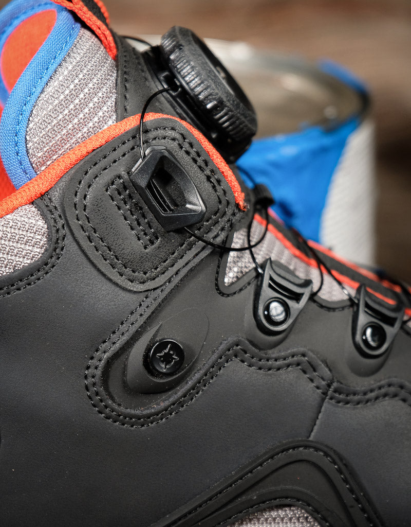 Close-up of a black high-top shoe with a dial closure system, featuring gray mesh panels, orange stitching, and blue accents.