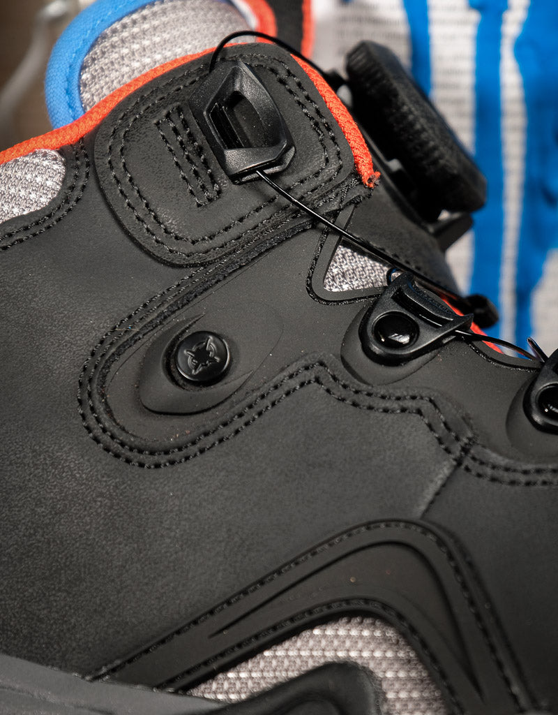 Close-up of a black high-top shoe with a dial closure system, gray mesh panels, and bright orange and blue accents.