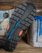 A close-up of a black rubber shoe sole with orange accents and the brand name 'DCSHOECUSA' printed in blue, resting on a rustic wooden surface with paint cans in the background.