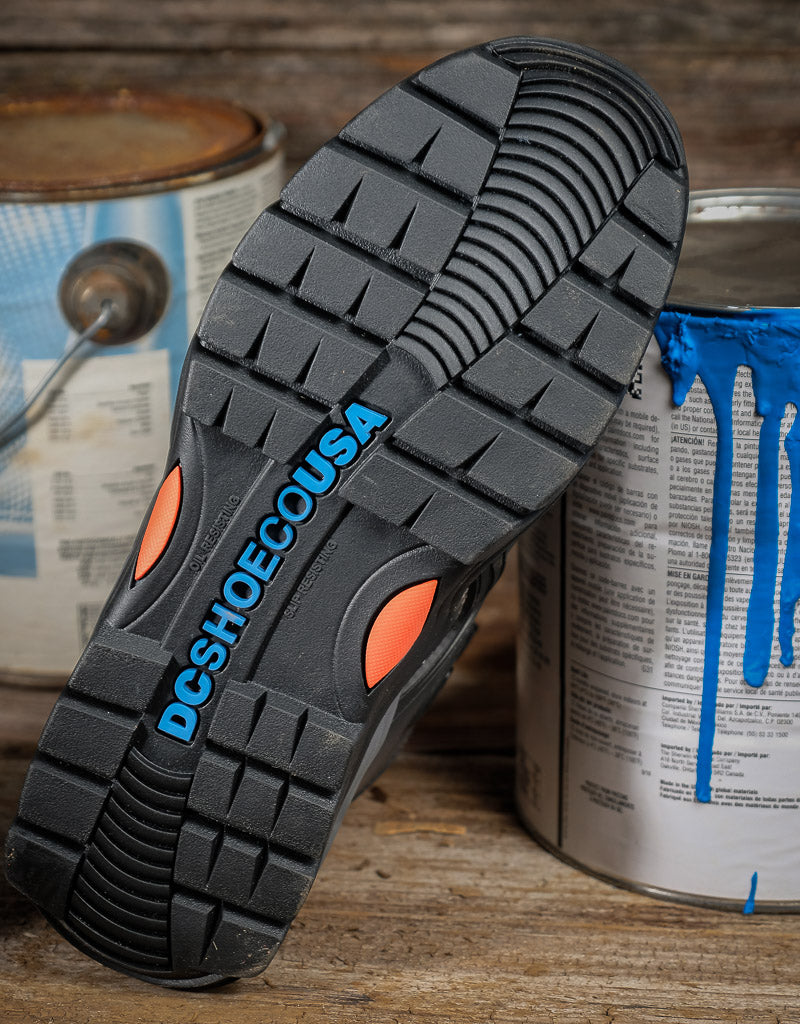 A close-up of a black rubber shoe sole with orange accents and the brand name 'DCSHOECUSA' printed in blue, resting on a rustic wooden surface with paint cans in the background.