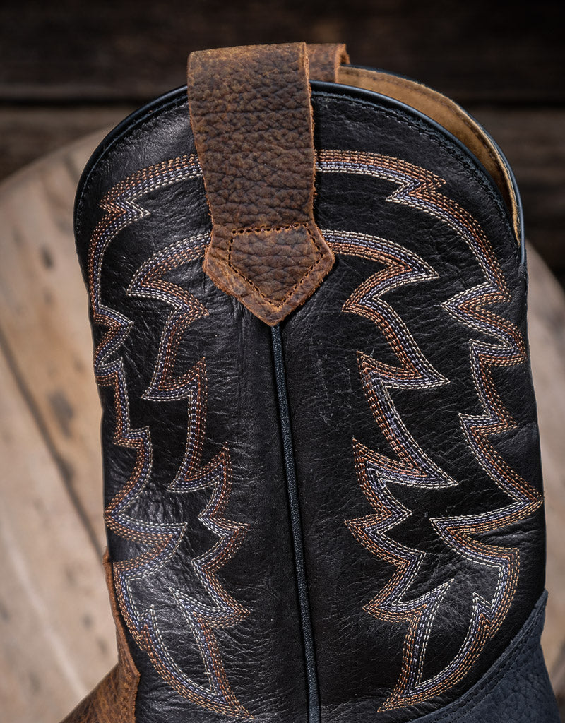 Close-up of a black leather boot shaft with intricate western stitching on a wooden surface.
