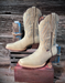 Pair of bone colored cowboy boots with intricate designs on a wooden stand against a rustic wooden background.