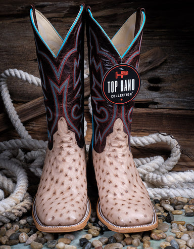 a pair of light colored ostrich western boots with maroon tops sit on top of rocks and rope with a background of wood