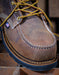 Close-up of a brown leather boot with a moc toe yellow laces on a wooden surface.