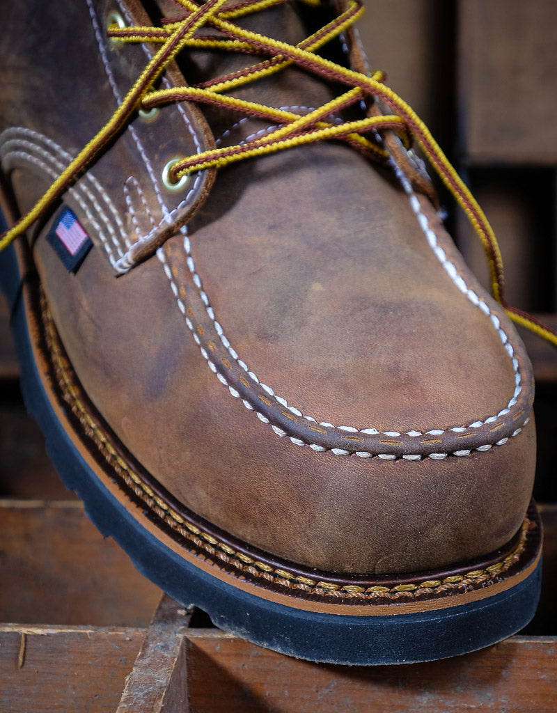 Close-up of a brown leather boot with a moc toe yellow laces on a wooden surface.