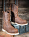 A pair of rugged brown leather work boots with thick off-white soles, displayed on a vintage blue-green trunk against a rustic wooden plank background.