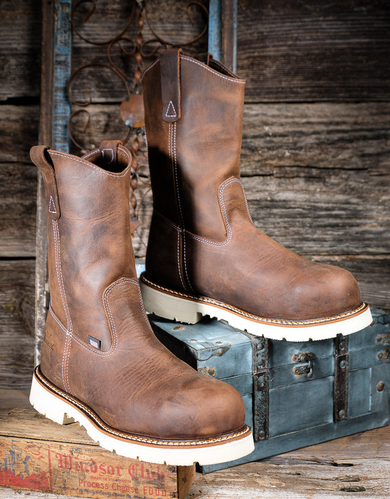 A pair of rugged brown leather work boots with thick off-white soles, displayed on a vintage blue-green trunk against a rustic wooden plank background.