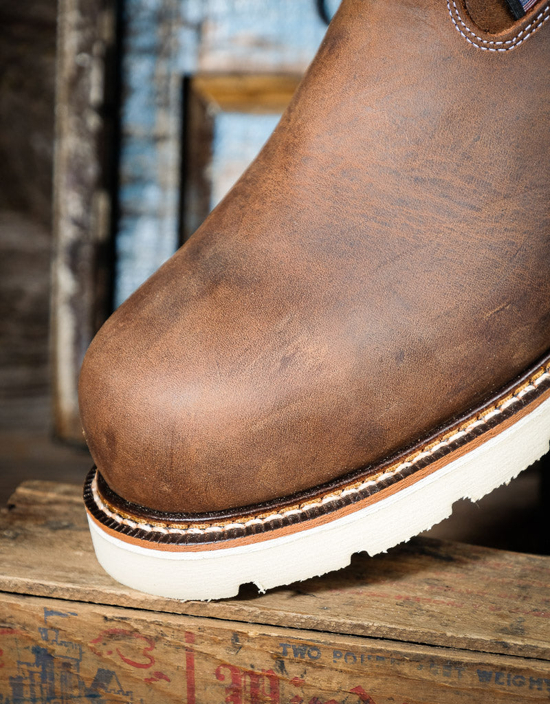 A close-up of a brown leather boot resting on a rustic wooden crate, showing the textured leather upper, decorative stitching, and a thick off-white sole.