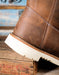 A close-up of a brown leather boot with white contrast stitching and a thick off-white sole, resting on a rustic wooden crate.