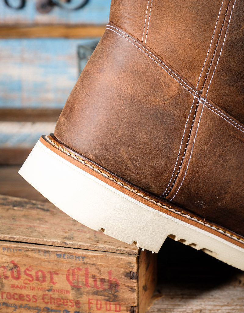 A close-up of a brown leather boot with white contrast stitching and a thick off-white sole, resting on a rustic wooden crate.
