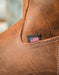 Close-up of a brown leather boot corner with white stitching and a small black fabric patch featuring an American flag.