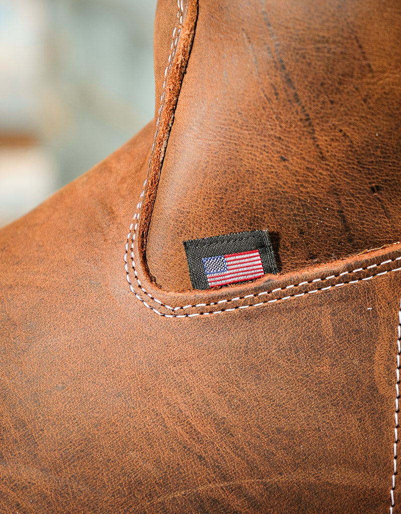 Close-up of a brown leather boot corner with white stitching and a small black fabric patch featuring an American flag.
