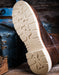 A close-up of a brown leather boot showing its beige treaded outsole, photographed on a rustic wooden surface with vintage blue and wood trunks in the background.