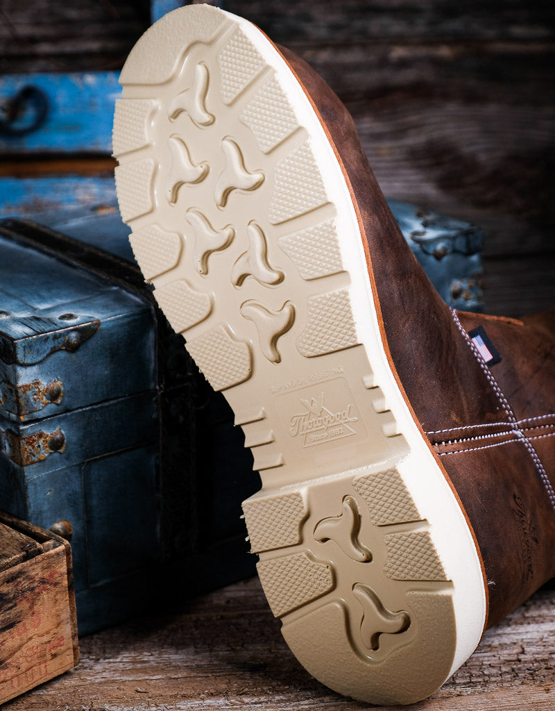 A close-up of a brown leather boot showing its beige treaded outsole, photographed on a rustic wooden surface with vintage blue and wood trunks in the background.