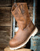 A brown distressed leather work boot with a thick white lug sole, displayed on a rustic wooden crate against a weathered wood and blue metal background.