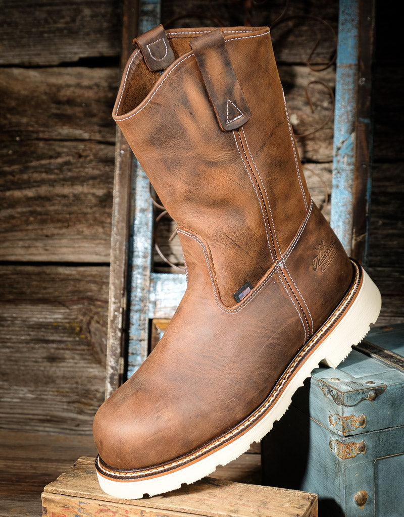 A brown distressed leather work boot with a thick white lug sole, displayed on a rustic wooden crate against a weathered wood and blue metal background.