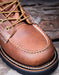 Close-up of a brown leather moc toe boot with yellow laces on a wooden surface