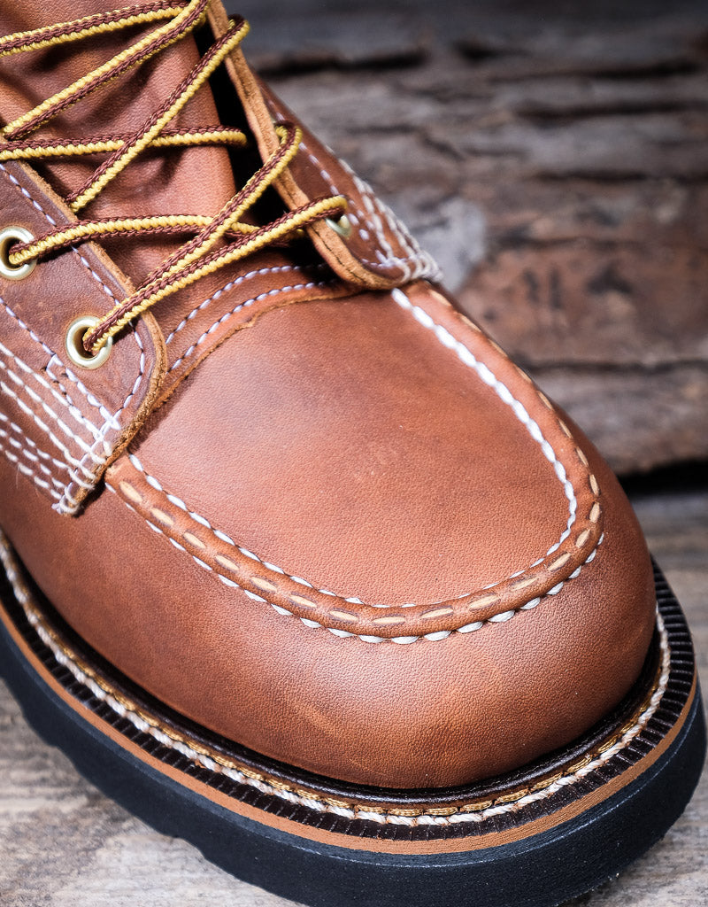 Close-up of a brown leather moc toe boot with yellow laces on a wooden surface