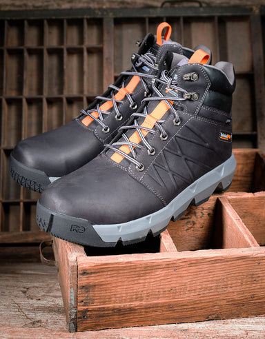 A pair of black high-top work boots with bright orange accents on the laces and heel pull tabs, displayed on a rustic wooden crate.