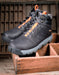 A pair of black high-top work boots with bright orange accents on the laces and heel pull tabs, displayed on a rustic wooden crate.