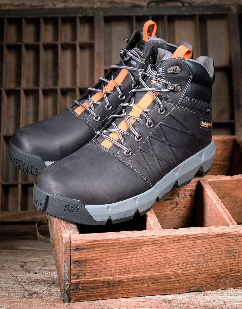 A pair of black high-top work boots with bright orange accents on the laces and heel pull tabs, displayed on a rustic wooden crate.