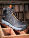 A single black high-top work boot with gray sole and bright orange accents, resting on a wooden crate in front of a rustic wooden grid background.