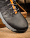 A close-up of a black protective work boot with gray laces featuring orange accents, resting on a rustic wooden surface.