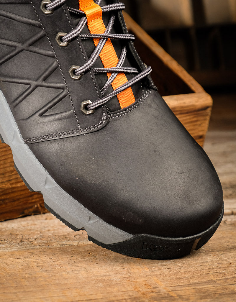 A close-up of a black protective work boot with gray laces featuring orange accents, resting on a rustic wooden surface.