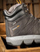 A close-up of a Timberland PRO mid-top work boot in black  leather with a light gray sole, resting on a rustic wooden surface.