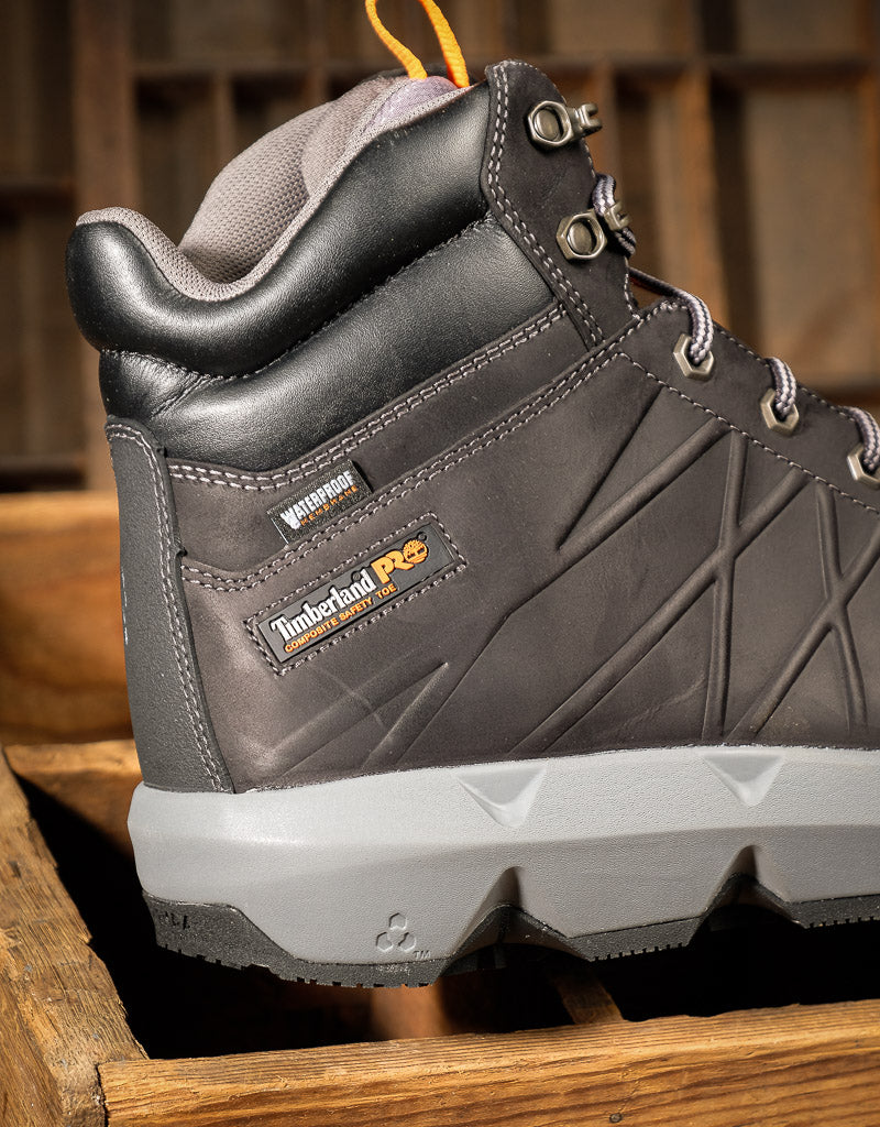 A close-up of a Timberland PRO mid-top work boot in black  leather with a light gray sole, resting on a rustic wooden surface.