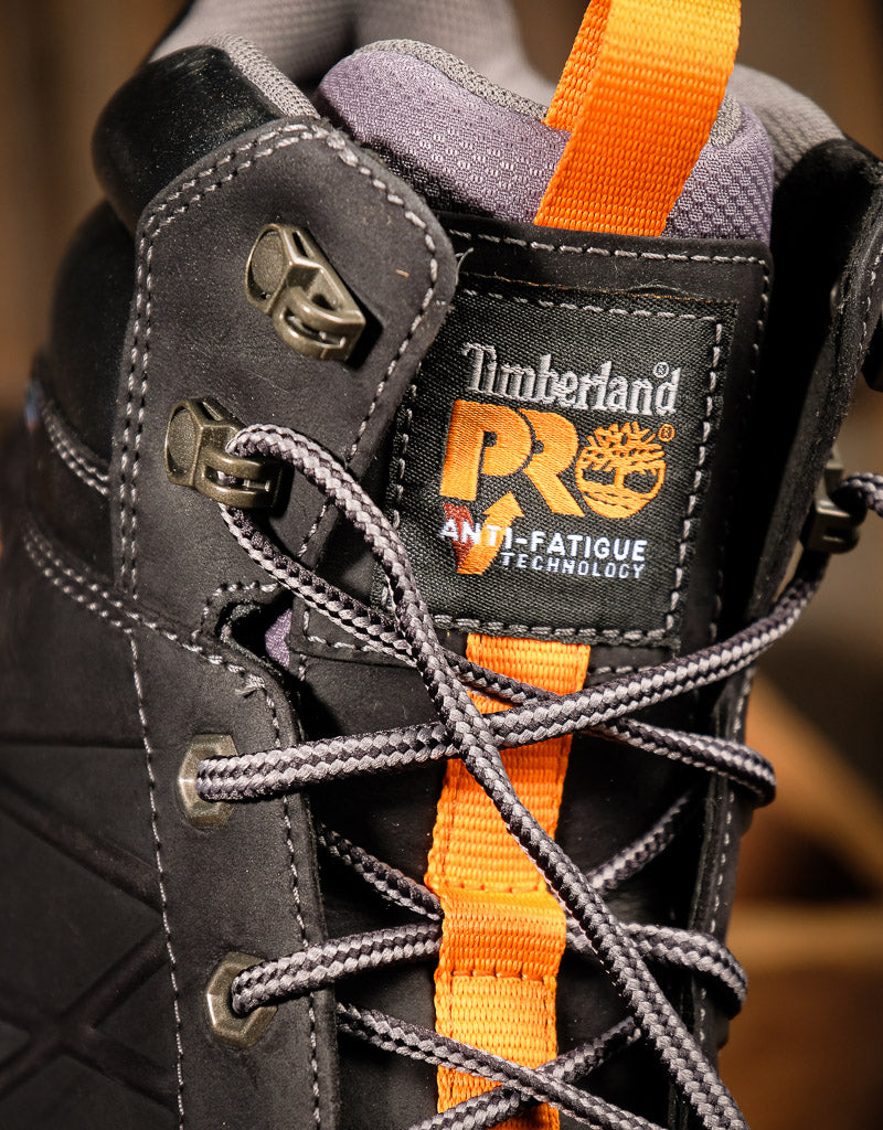 Close-up of a Timberland high-top work boot in  black with bright orange accents, including the tongue pull tab, lace loop, and branding patch.