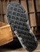 A close-up of a black rubber shoe sole with a deep tread pattern, resting on a rustic wooden surface with a wooden crate in the foreground.