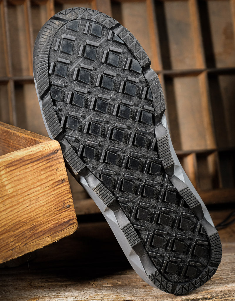 A close-up of a black rubber shoe sole with a deep tread pattern, resting on a rustic wooden surface with a wooden crate in the foreground.