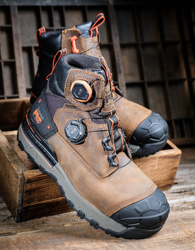 A pair of rugged work boots in brown and black with orange accents, displayed on wooden crates against a rustic dark wood background.